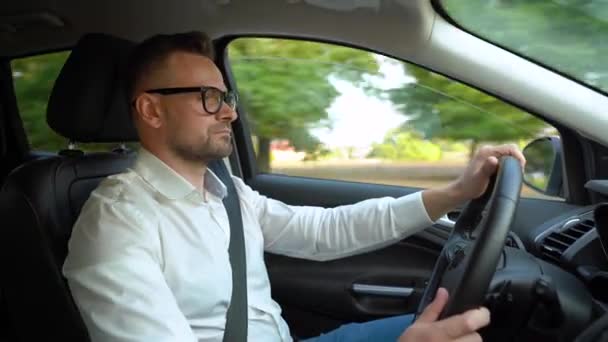 Homme barbu à lunettes et chemise blanche conduisant une voiture par temps ensoleillé. Vue latérale — Video