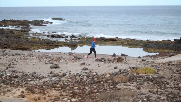 Una donna corre lungo la riva pietrosa dell'oceano. Stile di vita sano e attivo. Rallentatore — Video Stock