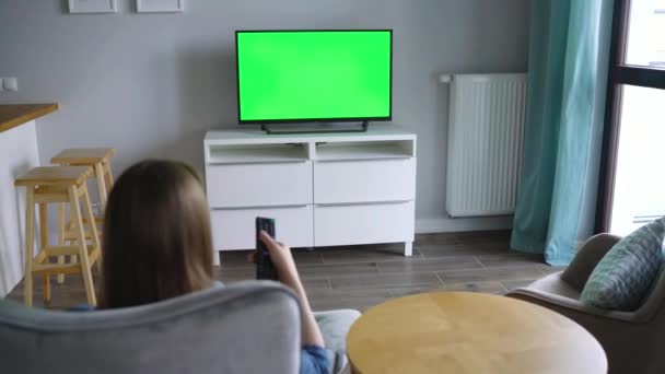 Donna è seduta su una sedia, guardando la TV con uno schermo verde, cambiando canale con un telecomando. Chiave cromatica. Interni — Video Stock