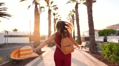 Gün batımında palmiye sokak boyunca uzanan büyük sarı şapkalı mutlu bir kadın. Yavaş çekim