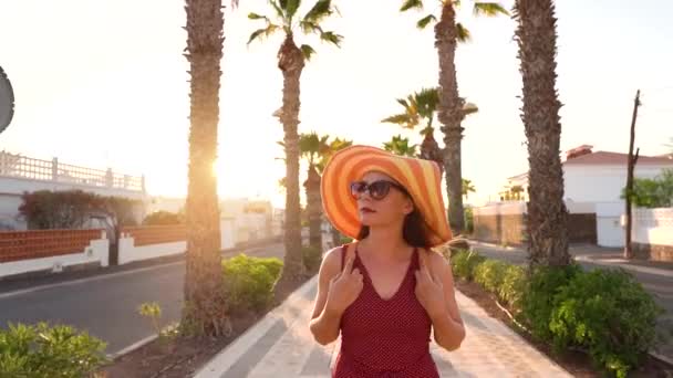 Glückliche Frau mit einem großen gelben Hut, die bei Sonnenuntergang eine Palmenallee entlanggeht — Stockvideo