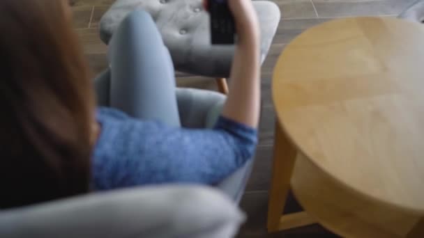 La mujer está sentada en una silla, viendo la televisión con una pantalla verde, cambiando los canales con un control remoto. Clave de croma. En interiores — Vídeos de Stock