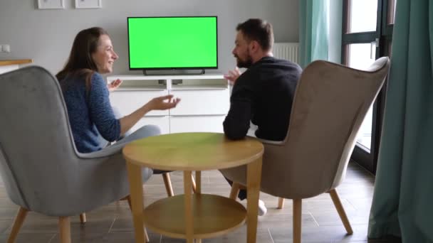 Homem e mulher estão sentados em cadeiras, assistindo TV com tela verde, discutindo o que viram e trocando de canais com um controle remoto. Vista traseira. Chave Chroma. Interior — Vídeo de Stock