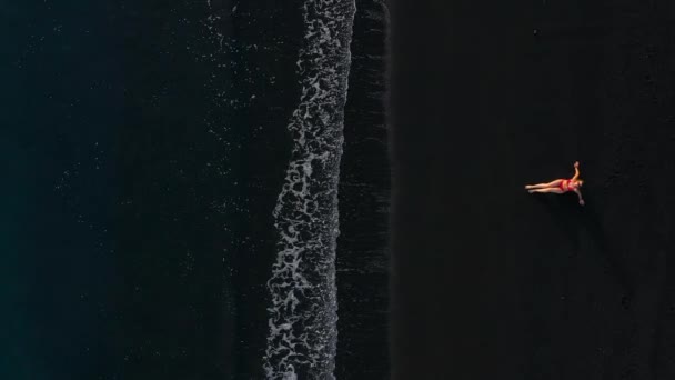 Top view egy lány piros fürdőruhában feküdt a fekete tengerparton a szörf vonalon. Tenerife sziget partja, Kanári-szigetek, Spanyolország. — Stock videók