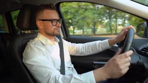 Бородатый мужчина в очках и белой рубашке водит машину в солнечную погоду и использует функцию автопилота во время вождения — стоковое видео