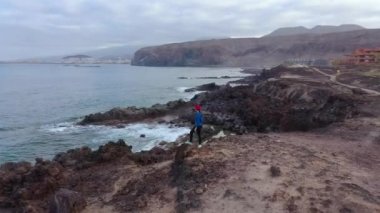 Spor giyimli bir kadının hava manzarası bir kayanın üzerinde duruyor ve Atlantik Okyanusu kıyısındaki bir koruma alanının manzarasının tadını çıkarıyor. Tenerife, Kanarya Adaları, İspanya