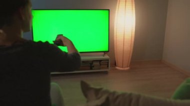 Kadın kanepede oturuyor, yeşil ekranlı televizyon izliyor, uzaktan kumandayla kanal değiştiriyor. Krom anahtar. Odanın köşesindeki kara kedi.