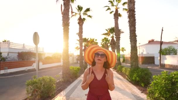 एक बड़ी पीली टोपी में खुश महिला सूर्यास्त पर एक पाम गली के साथ चल रही है। धीमी गति — स्टॉक वीडियो