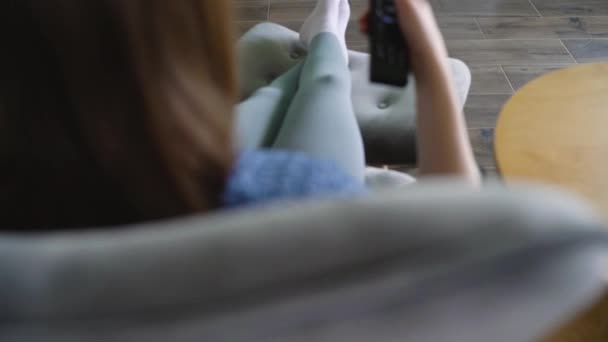 La mujer está sentada en una silla, viendo la televisión con una pantalla verde, cambiando los canales con un control remoto. Clave de croma. En interiores — Vídeos de Stock