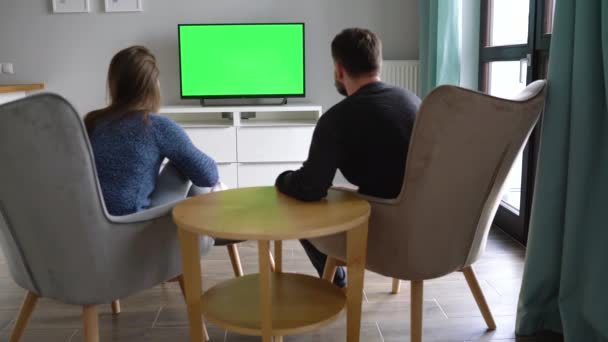 Man and woman are sitting in chairs, watching TV with a green screen, discuss what they saw and switching channels with a remote control. Back view. Chroma key. Indoors — Stock Video