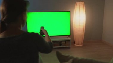 Kadın kanepede oturuyor, yeşil ekranlı televizyon izliyor, uzaktan kumandayla kanal değiştiriyor. Krom anahtar. Kapalı