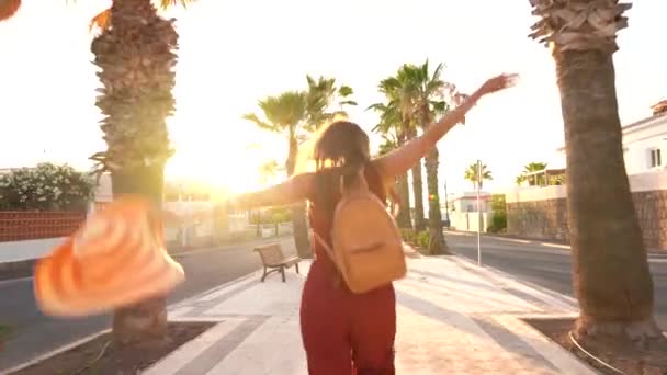 Ευτυχισμένη γυναίκα τρέχει χαρούμενα γερός κατά μήκος ενός σοκάκι παλάμη στο ηλιοβασίλεμα και κουνώντας ένα μεγάλο κίτρινο καπέλο — Αρχείο Βίντεο