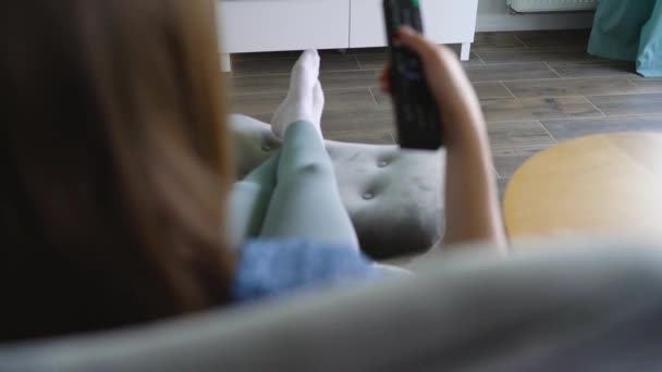 La mujer está sentada en una silla, viendo la televisión con una pantalla verde, cambiando los canales con un control remoto. Clave de croma. En interiores — Vídeos de Stock