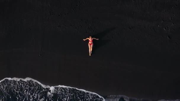 Draufsicht einer Frau im roten Badeanzug, die an einem schwarzen Strand an der Brandungslinie liegt. Küste der Insel Teneriffa, Kanarische Inseln, Spanien. — Stockvideo