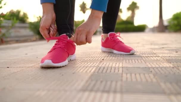 Primer plano de la mujer atando cordones de zapatos, inserta auriculares en las orejas y continúa corriendo a lo largo de la avenida de la palma al atardecer — Vídeos de Stock