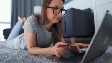 Kadın yerde yatıyor ve bir kredi kartı ve dizüstü bilgisayar kullanarak online alışveriş yapıyor. İnternetten alışveriş, yaşam tarzı teknolojisi.