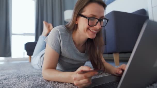 Mujer está tirado en el suelo y y hace una compra en línea con una tarjeta de crédito y un ordenador portátil. Ella es feliz y habla con alguien detrás de escena. Compras en línea, tecnología de estilo de vida — Vídeo de stock