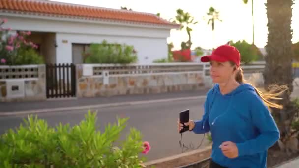 Woman with headphones and smartphone runs down the street along the palm avenue at sunset. Healthy active lifestyle — Stockvideo