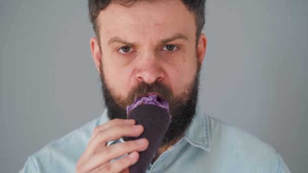 Close-up van een bebaarde man die paars ijs eet in een zwarte wafelbeker op een grijze achtergrond — Stockvideo