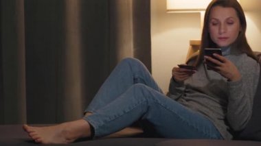 Sıcak bir odada kanepede uzanmış kredi kartı ve akıllı telefonuyla internetten alışveriş yapan bir kadın. İnternetten alışveriş, yaşam tarzı teknolojisi..