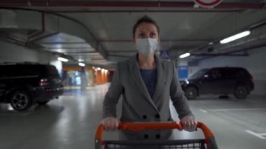 Tıbbi maskeli bir kadın bir market arabasıyla yeraltı otoparkından geçiyor. Covid-19 Coronavirus Pademia sırasında satın alma