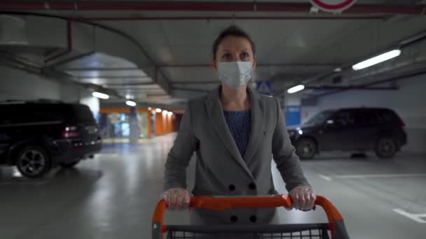 Kvinna i en medicinsk mask går med en matvagn genom en underjordisk parkeringsplats. Inköp under Covid-19 coronavirus pademia — Stockvideo