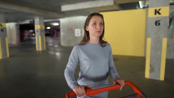 Kvinna går igenom en underjordisk parkeringsplats med en kundvagn — Stockvideo