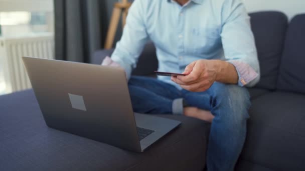 Hombre sentado en un sofá acogedor y hace el pago en línea con tarjeta de crédito. Servicio de pago en línea . — Vídeos de Stock
