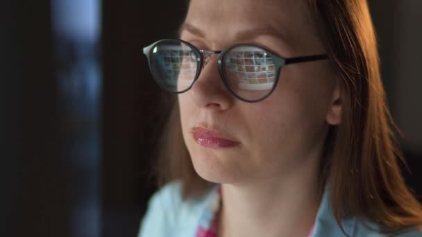 Een vrouw met een bril kijkt op de monitor en surft op internet. Het beeldscherm wordt gereflecteerd in de bril. Werk 's nachts. Binnenlandse Zaken. Afstandswerk — Stockvideo