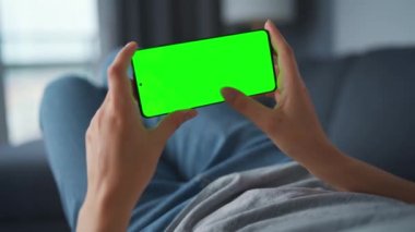 Evde bir kadın kanepeye uzanıyor ve yatay manzara modunda yeşil maket ekranlı akıllı telefon kullanıyor. İnternette gezinen, içerik izleyen, videolar, bloglar. POV.