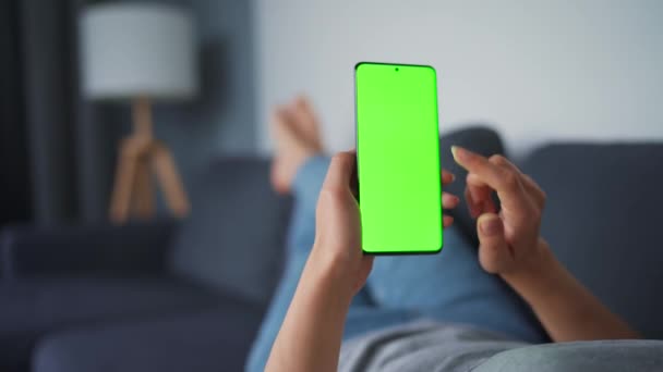 Kvinna hemma ligger på en soffa och använder smartphone med grön mock-up skärm i vertikalt läge. Flicka surfa på Internet, titta på innehåll, videor, bloggar. Födelsedatum. — Stockvideo