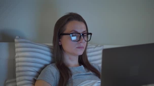 Frau mit Brille arbeitet am Laptop, während sie nachts im Bett liegt. Konzept für Internetsüchtige oder Schlaflosigkeit. — Stockvideo