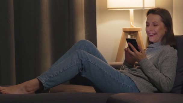 Femme souriante couchée sur le canapé dans une chambre confortable et utilisant un smartphone pour surfer sur Internet le soir. Elle rit de ce qu'elle voit. Relaxation et technologie lifestyle. — Video