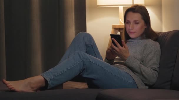 Vrouw liggend op de bank in een gezellige kamer en 's avonds met smartphone surfen op internet. Ontspanning en lifestyle technologie. — Stockvideo