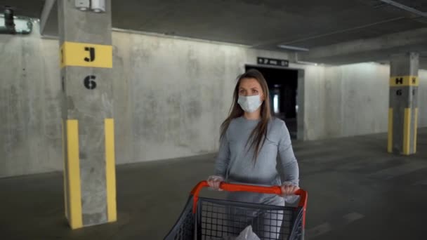 医療マスクの女性は地下駐車場を通って食料品店のカートで歩く。Covid-19コロナウイルスアカデミア期間中の購入 — ストック動画