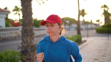 Kulaklıklı bir kadın gün batımında Palm Bulvarı boyunca koşuyor. Sağlıklı aktif yaşam tarzı. Yavaş çekim