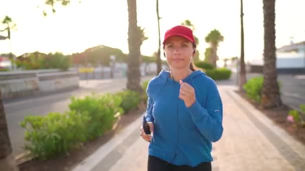 Femme avec casque et smartphone court dans la rue le long de l'avenue de la paume au coucher du soleil. Mode de vie sain et actif — Video