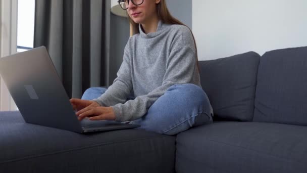 O femeie care stă pe o canapea confortabilă și lucrează la un laptop. Conceptul de muncă la distanță . — Videoclip de stoc
