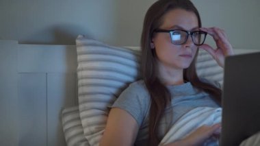Gözlüklü kadın geceleri yatakta yatarken dizüstü bilgisayarda çalışıyor. İnternet bağımlısı veya uykusuzluk konsepti.