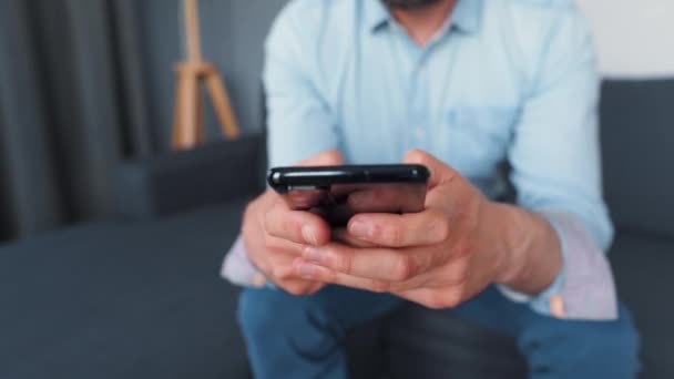 Homem vestido formalmente senta-se em um sofá e usando smartphone para mensagem de texto. Close-up — Vídeo de Stock