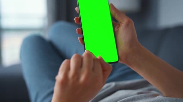 Женщина дома лежит на диване и использует смартфон с зеленым макетом экрана в вертикальном режиме. Девушка просматривает Интернет, смотрит контент, видео, блоги. POV. — стоковое видео