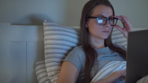 Mujer con gafas trabajando en el portátil mientras está acostada en la cama por la noche. Concepto de adicto a Internet o insomnio . — Vídeos de Stock