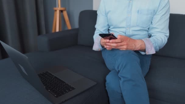 Mann sitzt auf einem gemütlichen Sofa und arbeitet an Laptop und Smartphone. Konzept der Fernarbeit. — Stockvideo