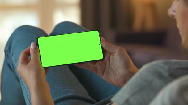 Mujer en casa acostada en un sofá y usando un smartphone con pantalla verde simulada en modo horizontal. Chica navegando por Internet, viendo contenido, videos, blogs. POV. — Vídeo de stock