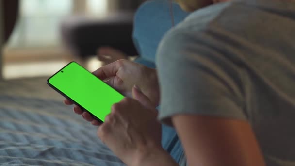 自宅の女性はソファに横になり、垂直モードで緑色のモックアップ画面を持つスマートフォンを使用しています。女の子はインターネットを閲覧し、コンテンツ、ビデオ、ブログを見ています。POV. — ストック動画
