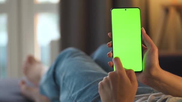 Mujer en casa acostada en un sofá y usando un smartphone con pantalla verde en modo vertical. Chica navegando por Internet, viendo contenido, videos, blogs. POV. — Vídeo de stock
