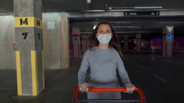 Женщина в медицинской маске ходит с тележкой с продуктами через подземную парковку. Покупка во время коронавирусной падемии Covid-19 — стоковое видео