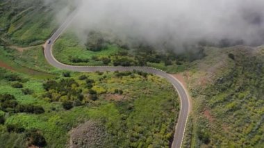 Yeşil bitki örtüsüyle çevrili bir dağ yolunda bulutların üzerinde uçuyorlar. Yolda giden mavi araba.