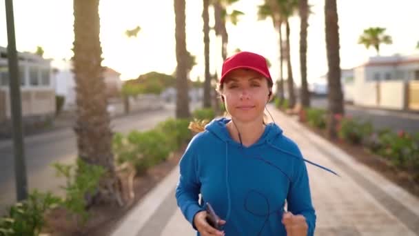 Kvinna med hörlurar och smartphone springer nerför gatan längs palmavenyn vid solnedgången. En hälsosam aktiv livsstil. Långsamma rörelser — Stockvideo
