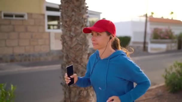 Mujer con auriculares y smartphone corre por la calle a lo largo de la avenida Palm al atardecer. Vida activa saludable. Movimiento lento — Vídeo de stock